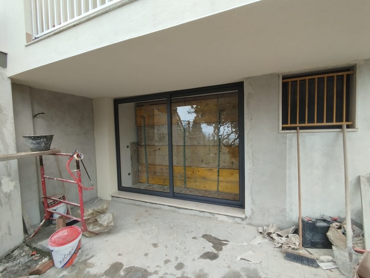 Portafinestra scorrevole con telaio scuro su facciata grezza in cantiere, tra secchi, carriola rossa e scope