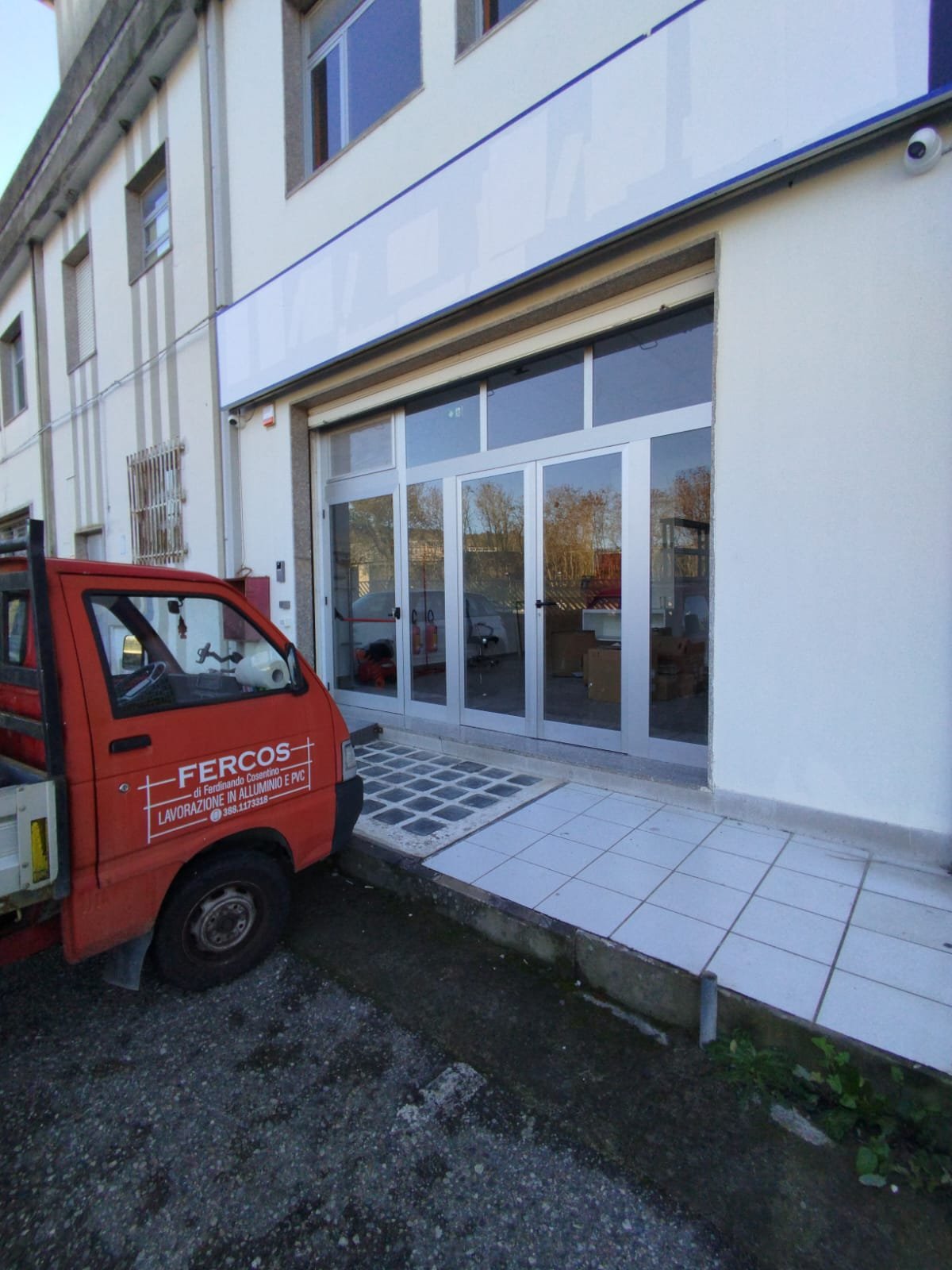 Furgoncino rosso con scritta FERCOS Lavorazione in alluminio e PVC parcheggiato davanti a un ingresso vetrato